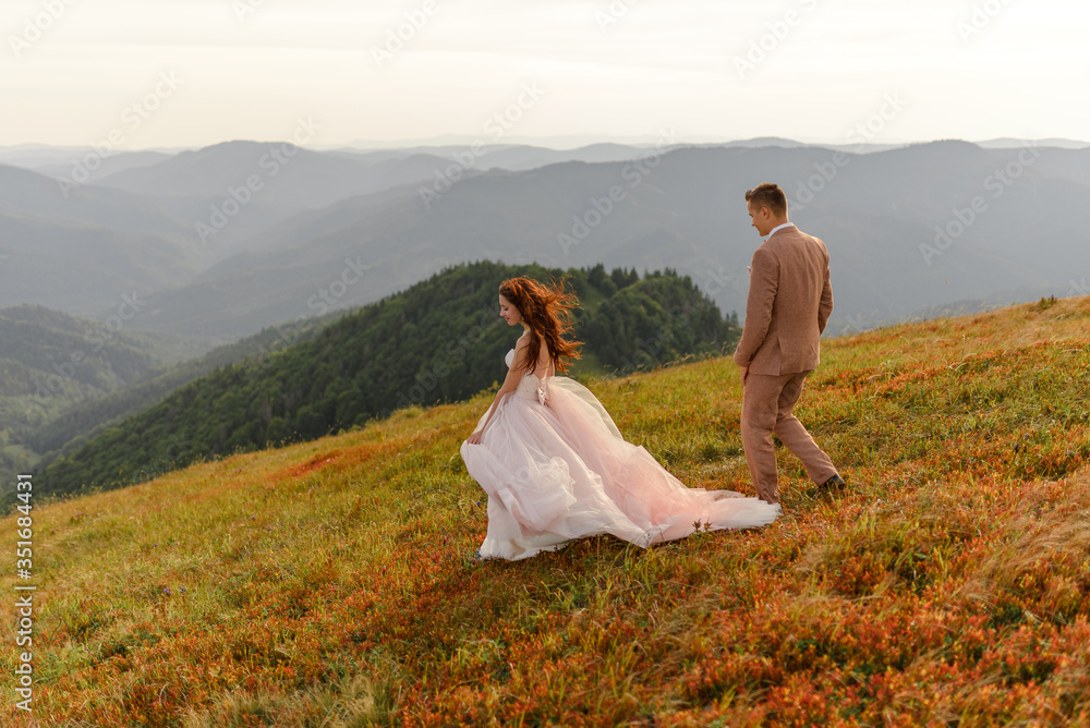 The bride and groom go next to each other. Sunset. Wedding photo on a background of autumn mountains. A strong wind inflates hair and dress.