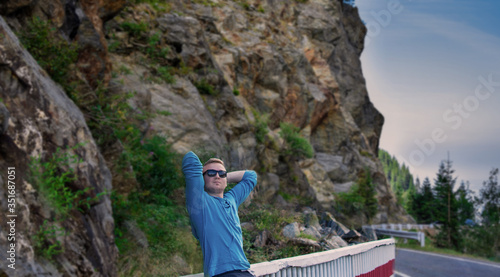 Young man in the middle of highway and looking away, enjoying fresh air near beautifull mountain. Portrait of traveler with sunglasses walking down the road.