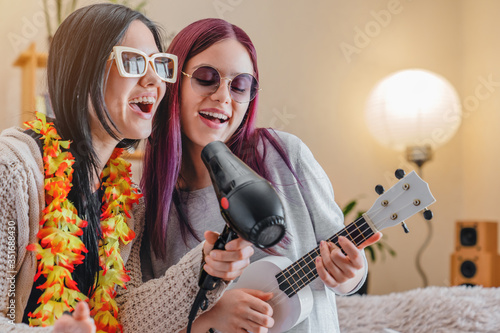 Young joyful girls in sunglasses dancing and singing with hairdryer and ukulele at home