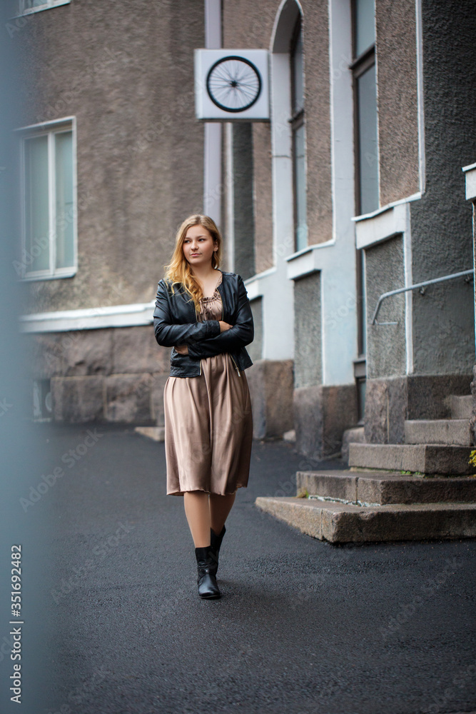 Fototapeta premium Young fashion blond woman walking on city street Stylish female model in black leather jacket and drees