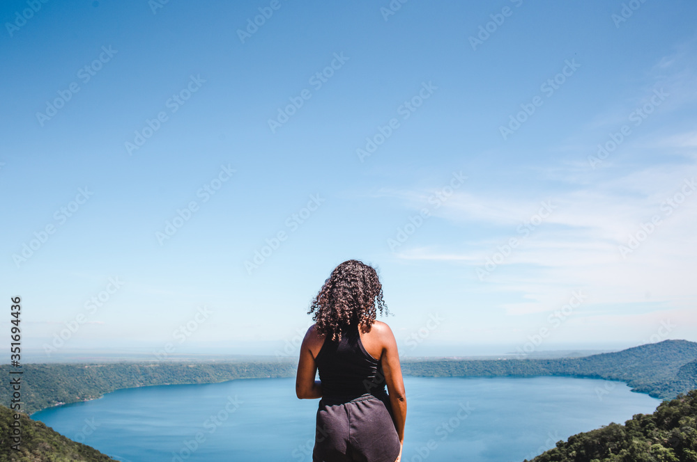 Mixed race millennial backpacker girl looks out form aerial viewpoint over Laguna de Apoyo, a crater lake near Masaya, Nicaragua