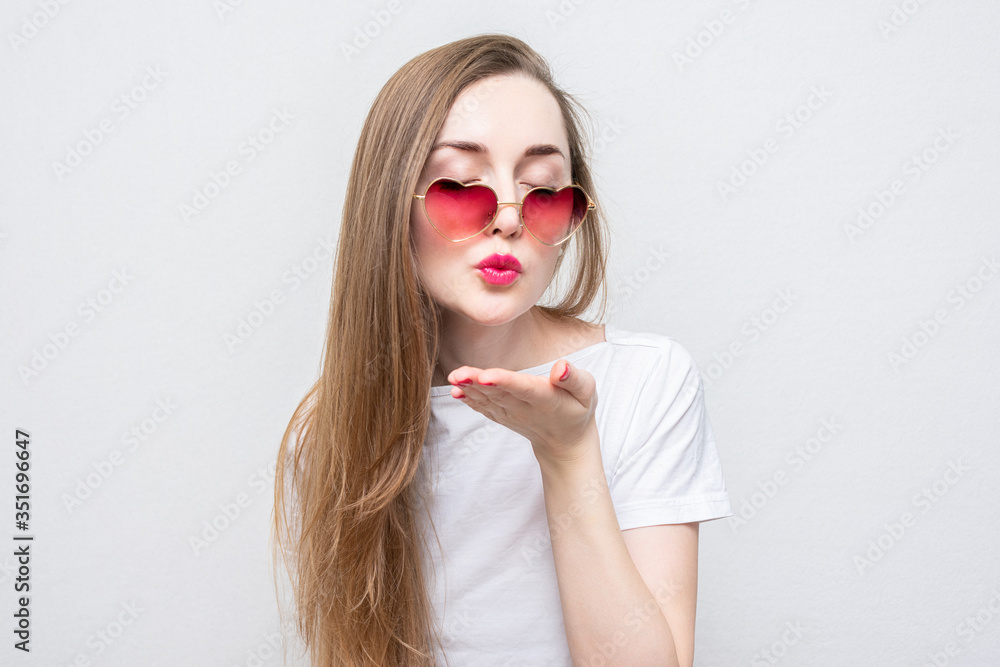 Young woman sends air kiss, girl heart-shaped glasses, white background, copy space Young woman sends air kiss, girl heart-shaped glasses, white background, copy space