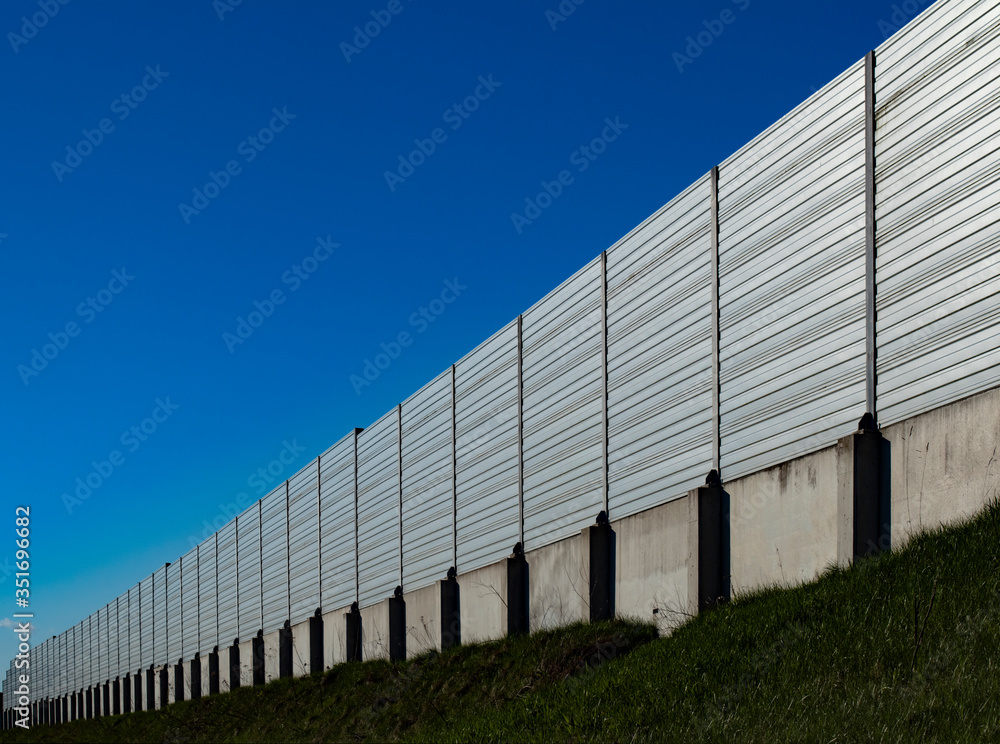 A concrete and metal panel sound barrier at the side of the road, outside perspective view with grass and blue clear sky A concrete and metal panel sound barrier at the side of the road, outside perspective view with grass and blue clear sky
