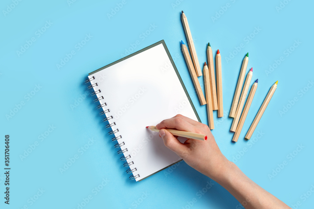 Hand with a pencil over a notebook on a blue background. Nearby are colored pencils. Mockup, flat layout. Hand with a pencil over a notebook on a blue background. Nearby are colored pencils. Mockup, flat layout.