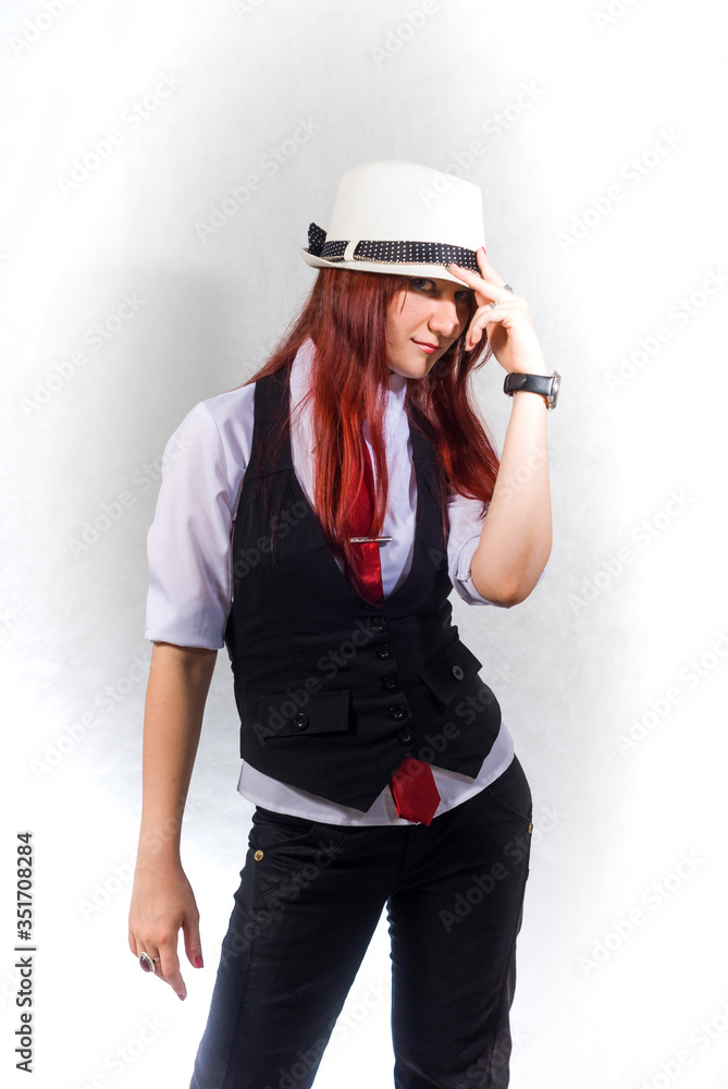 A beautiful young red-haired woman in a white blouse, black vest and red tie and a white hat on a white background, with her head down, looks out from under the brim of her hat. A beautiful young red-haired woman in a white blouse, black vest and red tie and a white hat on a white background, with her head down, looks out from under the brim of her hat.