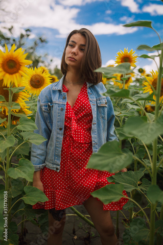 young beautiful caucasian girl in red dress in white dots stay in sunflower field 