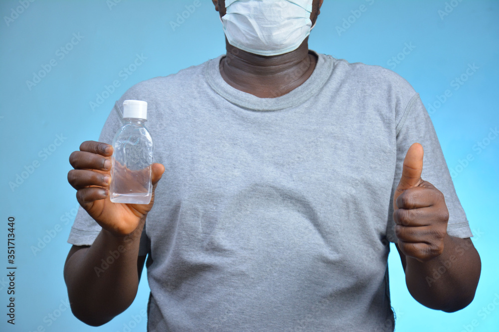 cropped image of a young african boy wearing a medical face mask and holding a bottle of a hand sanitizer with his thumbs up. African boy standing in front of a blue background. cropped image of a young african boy wearing a medical face mask and holding a bottle of a hand sanitizer with his thumbs up. African boy standing in front of a blue background.