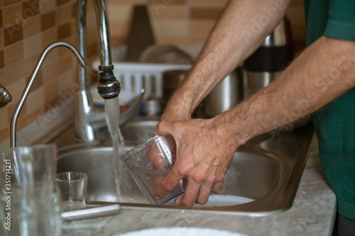 Obraz na plátně Men housework washes dishes in the sink, a dirty pan and plates under running water, the husband helps with the housework during quarantine of the coronavirus