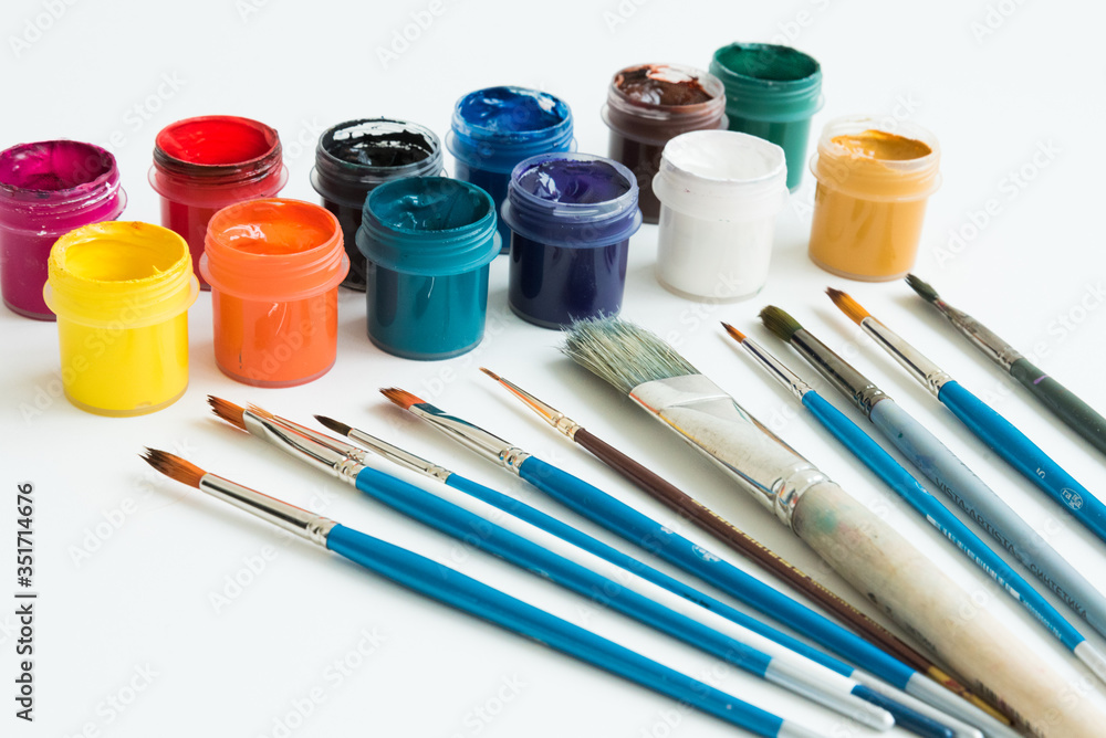 Jars of gouache paint in different colors. Next to it are brushes of various sizes. Artist's kit. Side view. White background. Hobbies and inspiration. Jars of gouache paint in different colors. Next to it are brushes of various sizes. Artist's kit. Side view. White background. Hobbies and inspiration.