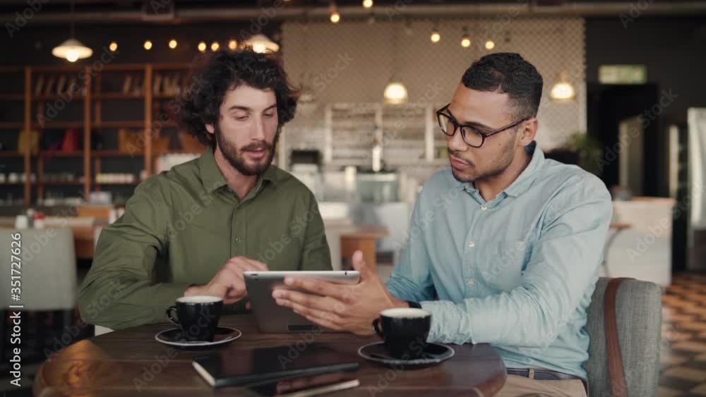 Multiethnic business man showing presentation to customer in cafe using digital tablet while drinking coffee - two men looking at a tablet Multiethnic business man showing presentation to customer in cafe using digital tablet while drinking coffee - two men looking at a tablet