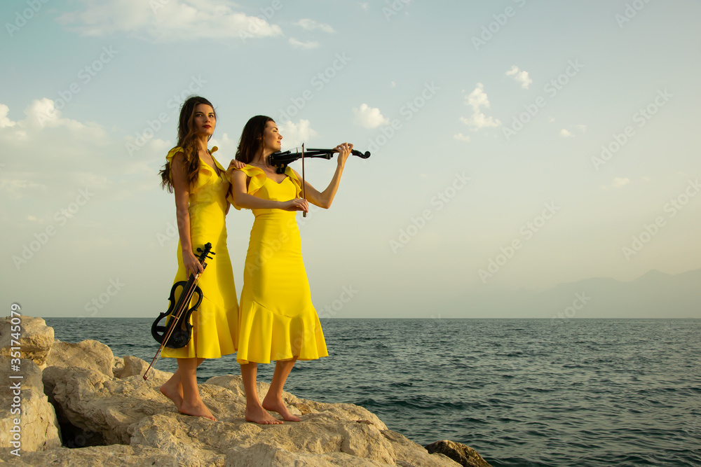 Fototapeta premium Two beautiful twin sisters violinists in yellow concert dresses are playing electric violins on rocks by the Mediterranean sea in Antalya, Turkey.
