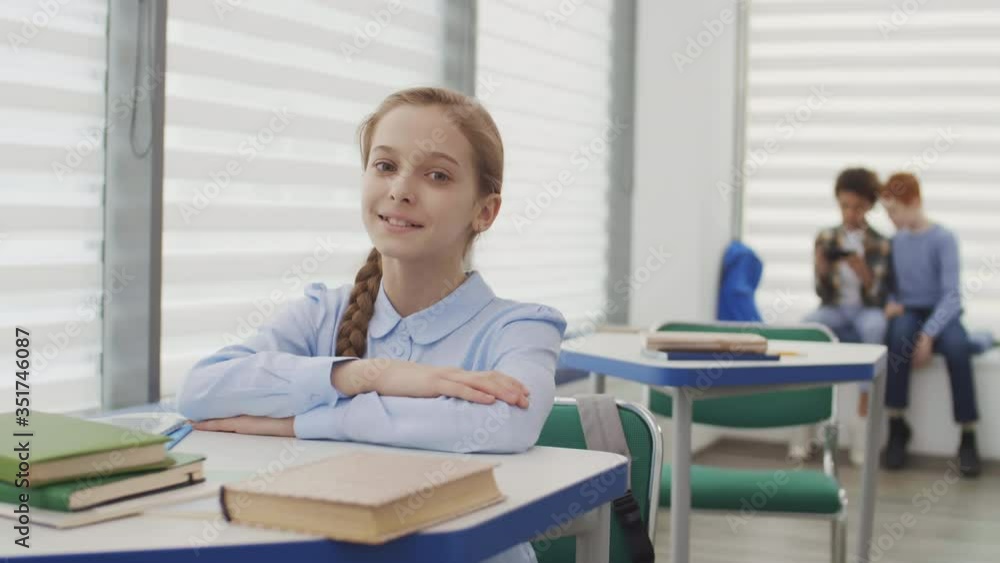 Medium shot portrait of blond-haired Caucasian girl sitting at desk in classroom and looking at camera while two boys sitting on background Medium shot portrait of blond-haired Caucasian girl sitting at desk in classroom and looking at camera while two boys sitting on background