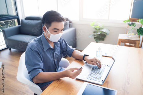 Asian man work from home ordering product online from ecommerce store, using mobile phone credit card payment transaction, wearing facemask protection from coronavirus covid-19 in quarantine isolation