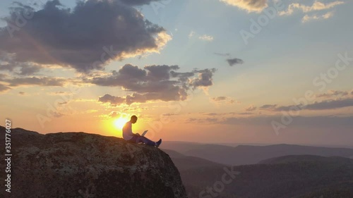 Aerial view of a young male freelancer working on a laptop sitting on a rock ledge in the mountains at sunset.