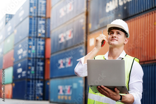Engineer or foreman use walkie-talkie and laptop. In order to move container and shipping. Safety helmet help prevent accident from various equipment in factory. Industry and Transportation concept