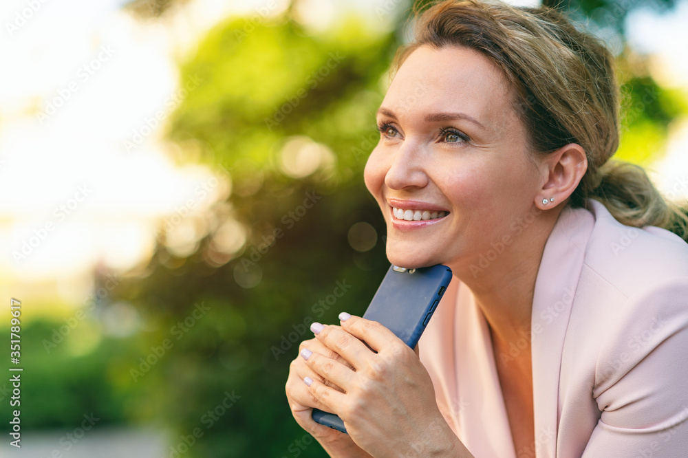 Close-up portrait of a smiling woman with smartphone on the street.  Happy businesswoman is using phone, outdoors. Cheerful businesswoman in a jacket with cell phone in park looking away.
