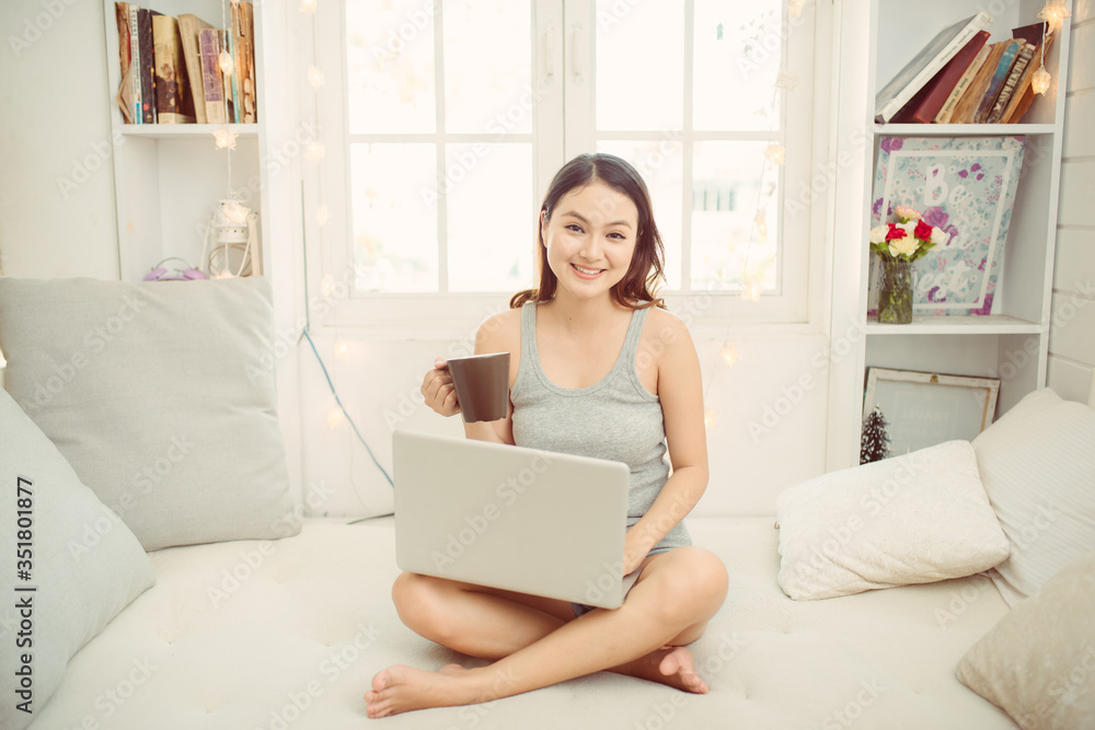 Happy beautiful woman working on a laptop and drinking coffee from a mug, while sitting on the bed in the house. Happy beautiful woman working on a laptop and drinking coffee from a mug, while sitting on the bed in the house.
