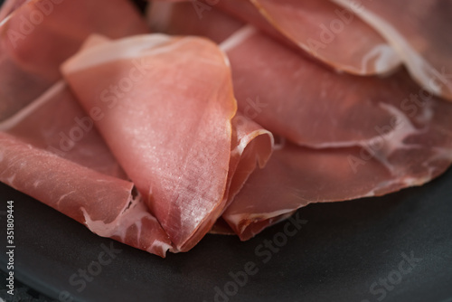 Thin italian prosciutto slices on black plate on conctere background closeup