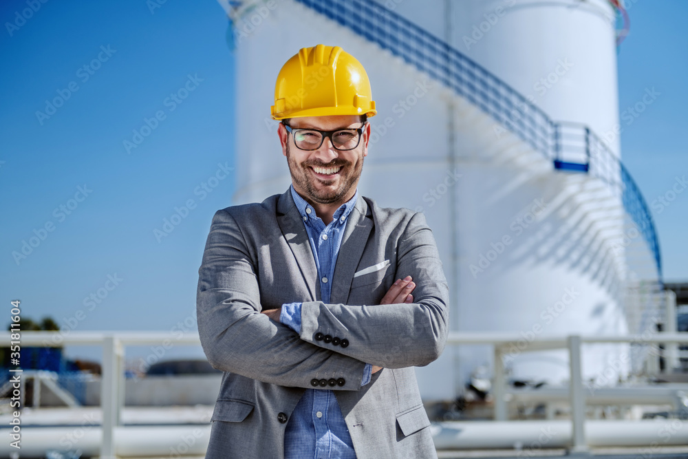 Young proud smiling successful businessman in suit with helmet on head standing with arms crossed. Refinery exterior. In background is tank with oil. Young proud smiling successful businessman in suit with helmet on head standing with arms crossed. Refinery exterior. In background is tank with oil.
