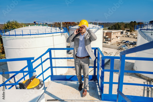 Full length of handsome elegant rich businessman having important call about oil selling. Refinery exterior. In background are oil tanks.