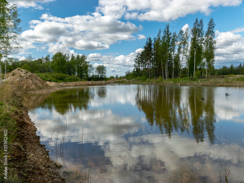Fototapeta Naklejka Na Ścianę i Meble -  a fish pond, in the water of which there are colorful cumulus clouds and trees, there are recently dug gravel piles on the shore of the pond, the first spring greenery in nature