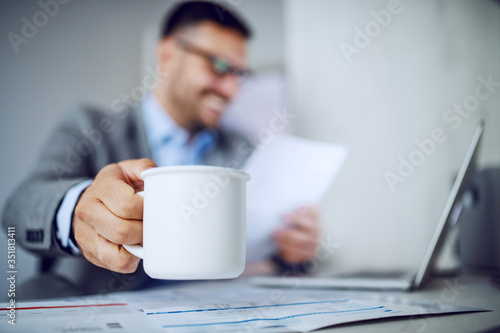 Beautiful caucasian classy smiling businessman in suit and with eyeglasses holding in one hand coffee and in other document and looking at it. Selective focus on mug with coffee. Office interior.