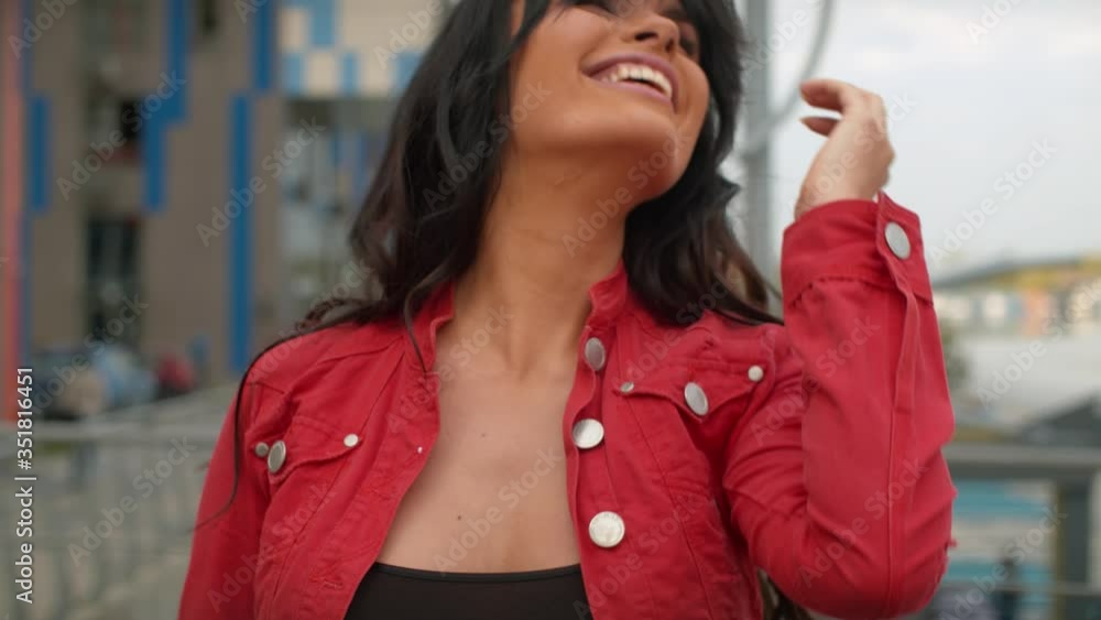 Close up portrait of smiling pretty woman with long beautiful hair standing next to a modern building. Attractive girl in red jacket in the city. Real people Close up portrait of smiling pretty woman with long beautiful hair standing next to a modern building. Attractive girl in red jacket in the city. Real people