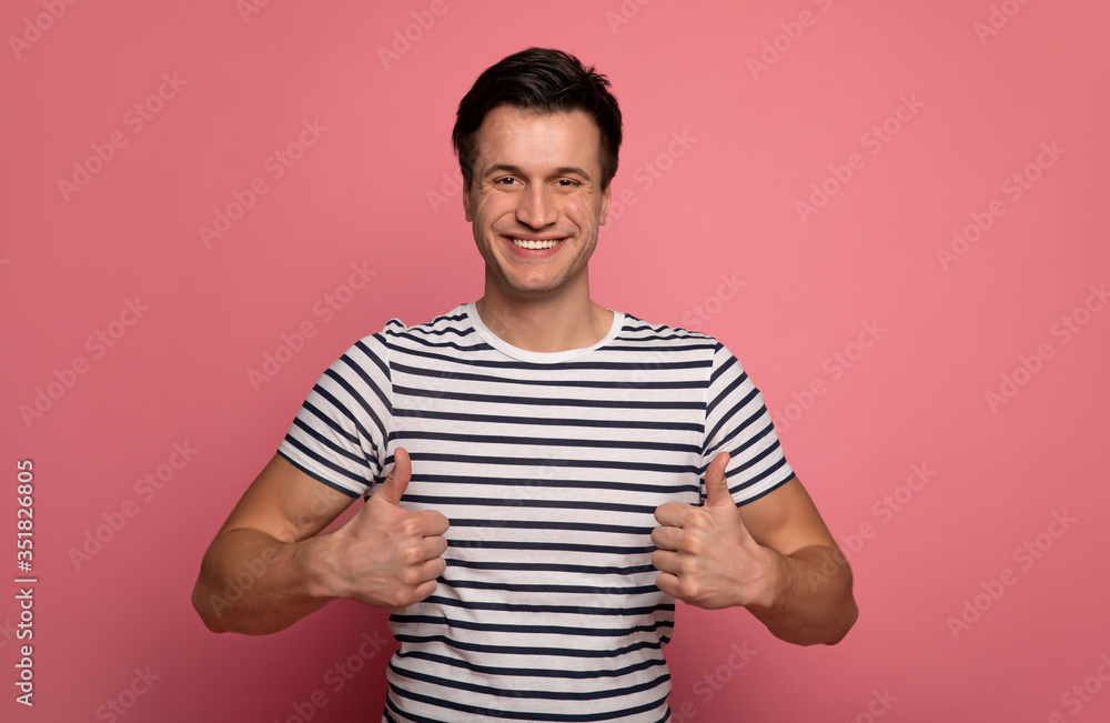 Positive attitude. Close-up photo of a young man in a striped t-shirt, who is looking in the camera, smiling and showing thumbs up. Positive attitude. Close-up photo of a young man in a striped t-shirt, who is looking in the camera, smiling and showing thumbs up.