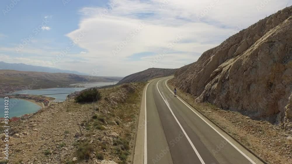 Aerial shot of man cycling on road by rock formation near sea, drone ascending over person riding on highway during sunny day - Pag, Croatia Aerial shot of man cycling on road by rock formation near sea, drone ascending over person riding on highway during sunny day - Pag, Croatia