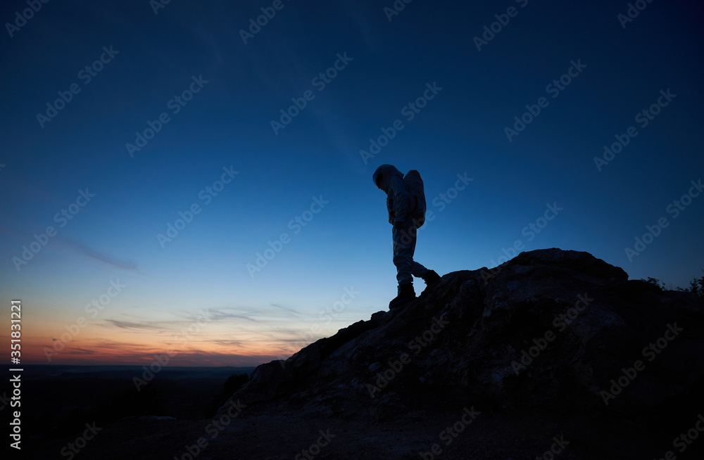 Beautiful view of space traveler on rocky mountain with majestic night sky on background. Silhouette of cosmonaut in space suit exploring new planet. Concept of space travel and twilight beauty.