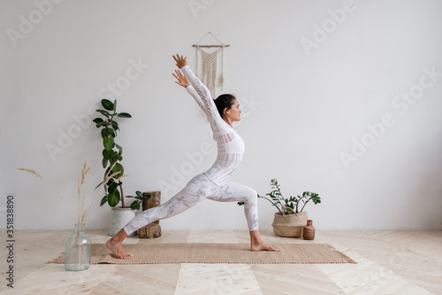 Side view of a slim pretty positive young brunette woman doing a virabhadrasana standing on the mat on the floor surrounded by houseplants on a white wall. Advertising space. Yoga and pilates photo