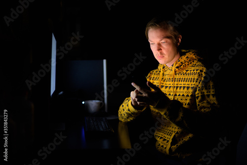 Young handsome man using phone while working overtime at home in the dark