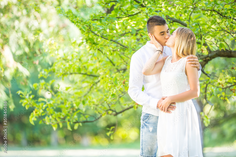 Young couple kissing under the green tree with summer nature background. Dressed in white dress, white shirt and jeans, holding hands Young couple kissing under the green tree with summer nature background. Dressed in white dress, white shirt and jeans, holding hands