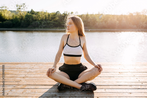 Young slim fit woman stretching before workout, sitting on a wooden platform by the lake, outdoors, in the morning.