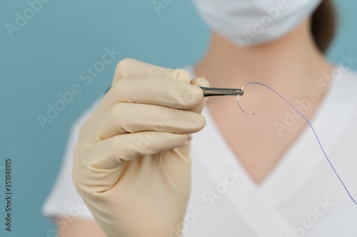 doctor holds a surgical thread with a needle in gloves with a mask on a blue background