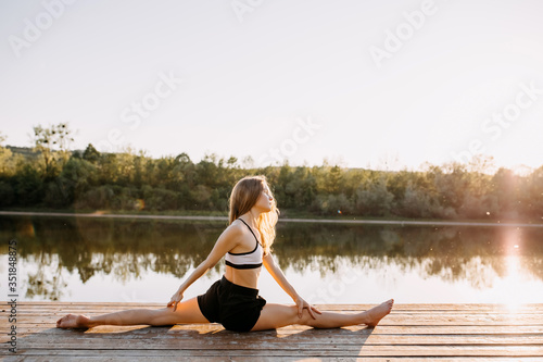 Young slim fit woman doing yoga exercise, twine pose, in the morning. Sportive girl outdoors, on a wooden platform by the lake.