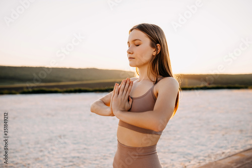 Young slim fit woman with hands to heart center, standing in yoga tree pose at the sunset, outdoors.