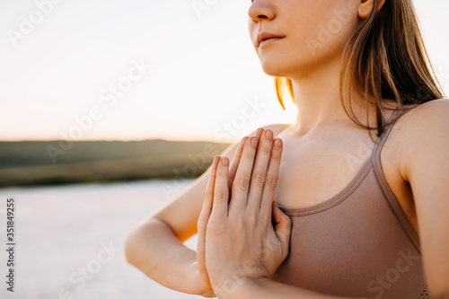 Close-up of a young woman with hands to heart center, standing in yoga pose at the sunset, outdoors.