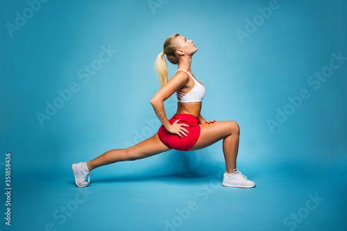 young fitness happy sexy blonde girl in red skirt and white top is doing yoga exercise and looking up with hands on hips on the blue wall background, sport concept, free space