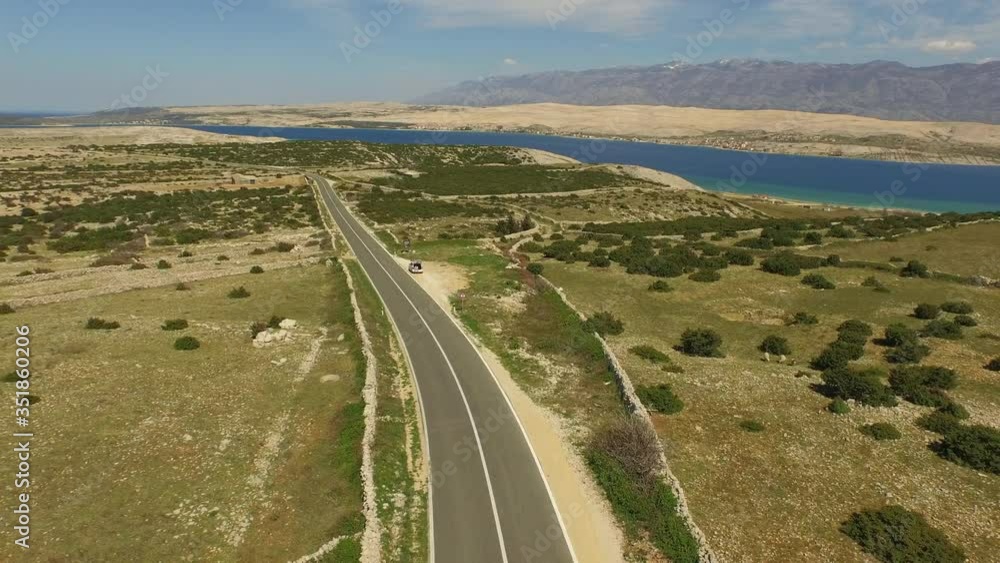Aerial shot of vehicles on highway amidst plants near sea, drone flying backward over landscape against sky on sunny day - Pag, Croatia Aerial shot of vehicles on highway amidst plants near sea, drone flying backward over landscape against sky on sunny day - Pag, Croatia