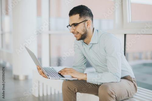 Happy intern sitting on a bench and working on a laptop in a bright office hall