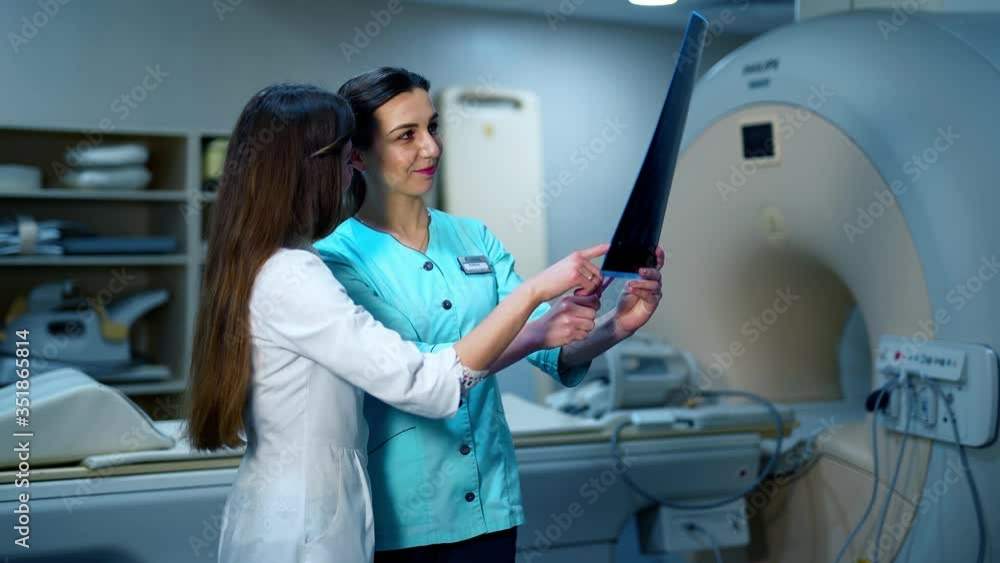 Medical workers discuss the x-ray photograph. Two female specialists in medical uniform examining x-ray photo on the background of new equipment in clinic. Medical workers discuss the x-ray photograph. Two female specialists in medical uniform examining x-ray photo on the background of new equipment in clinic.