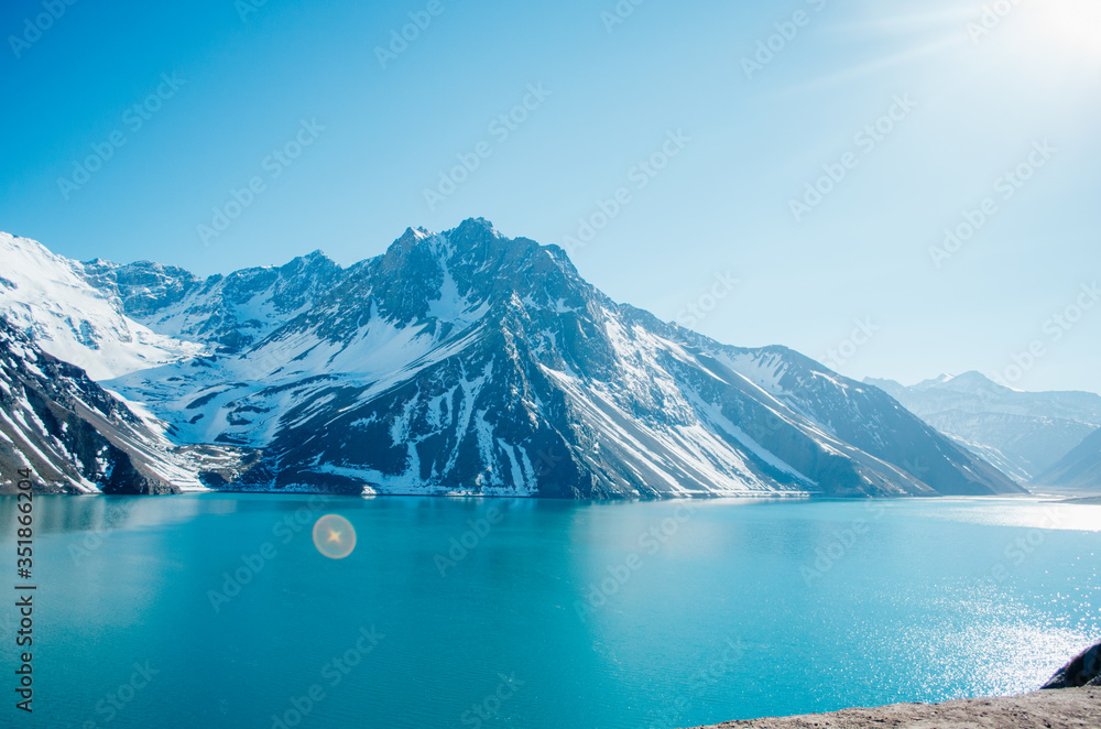 A dramatic panorama of El Yeso Dam with turquoise color and sun flares reflecting in the water in the Andes Mountains with snow on the peaks and rock parts of the mountain exposed.