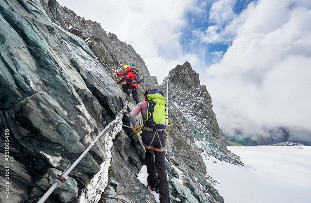 Back view of rock climbers with backpacks using fixed rope while ascending high rocky mountain. Male mountaineers climbing natural rock formation and trying to reach mountaintop. Back view of rock climbers with backpacks using fixed rope while ascending high rocky mountain. Male mountaineers climbing natural rock formation and trying to reach mountaintop.