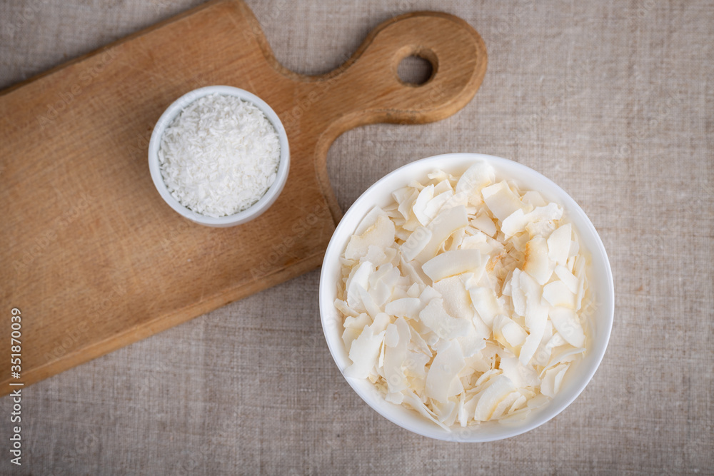 Coconut chips and shavings in a bowl on a cloth tablecloth and cutting board.Top view. Coconut chips and shavings in a bowl on a cloth tablecloth and cutting board.Top view.
