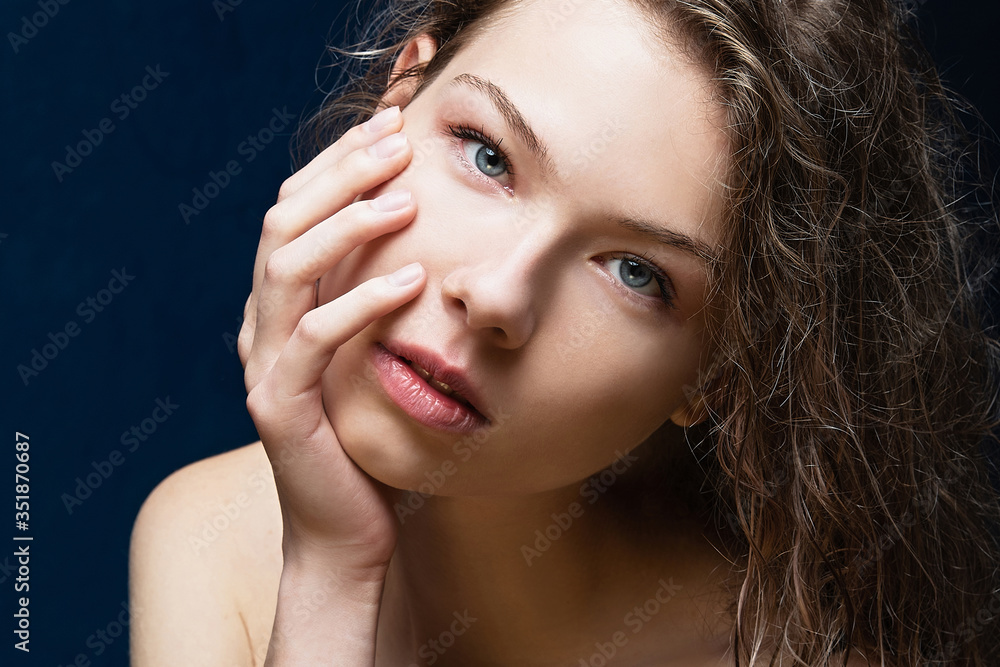 Fototapeta premium portrait of a young girl with long curly wet hair, close-up, fair skin, holds her face, looks forward, bare shoulders, smooth skin, blue eyes, attractive young girl