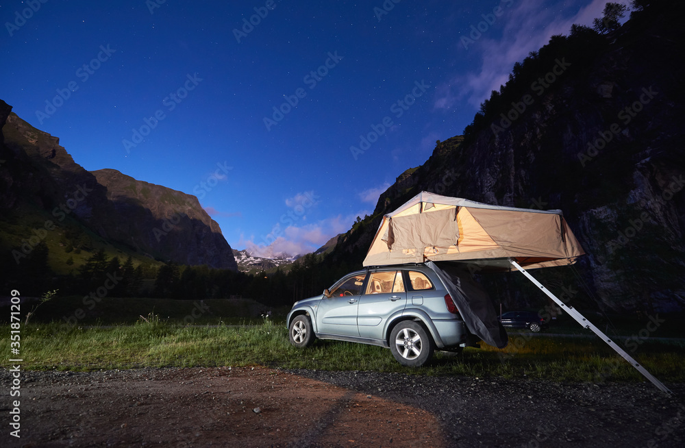 Beautiful view of blue night sky over mountains and automobile with camping  tent on rooftop. Camping evening scene with silver car and majestic hills  on background. Concept of travelling, camping. Stock Photo