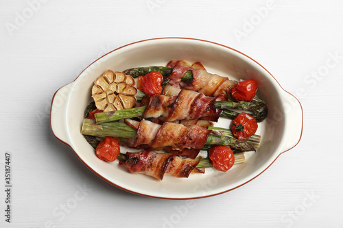 Oven baked asparagus wrapped with bacon in ceramic dish on white wooden table, top view