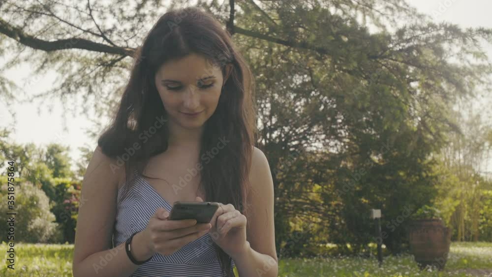 Dolly motion shot of Cute young woman with mobile phone typing browsing online on social media, relaxing resting outdoor sitting in park grass, sunny day in spring summer season. blurred background Dolly motion shot of Cute young woman with mobile phone typing browsing online on social media, relaxing resting outdoor sitting in park grass, sunny day in spring summer season. blurred background