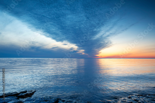 Cloudy summer sunset reflecting in ocean with endless horizon and deep blue ocean, silhouette of boulders laying in the foreground in shallow water at island of Gotland, Sweden